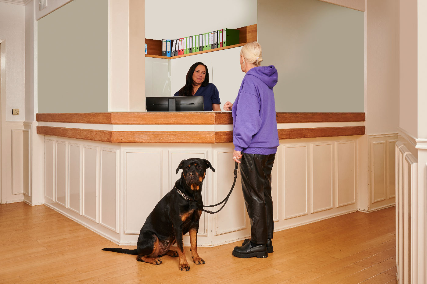 Medizinisches-Kleintierzentrum-Delmenhorst_vivus_kosten Eine Frau in einem lila Kapuzenpullover steht an einem Empfangstresen. Ein schwarz-brauner Hund sitzt an der Leine. Hinter dem Tresen sitzt eine Rezeptionistin.