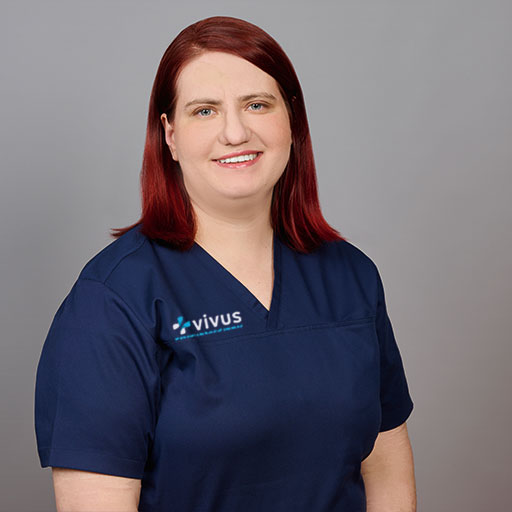 Marta_Dedko_vivus Eine Frau in einem marineblauen medizinischen OP-Kittel mit einem „Vivus“-Logo steht vor einem schlichten grauen Hintergrund und lächelt in die Kamera.